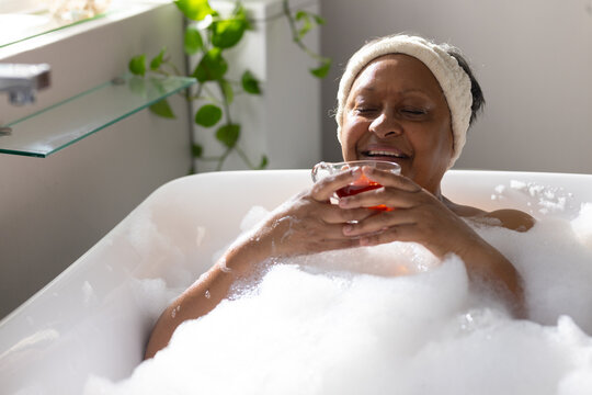Senior African American woman reclining in bath with foam holding glass with amber liquid