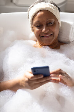 Senior African American woman reclining on towel in tub with foam using cell wearing white headband