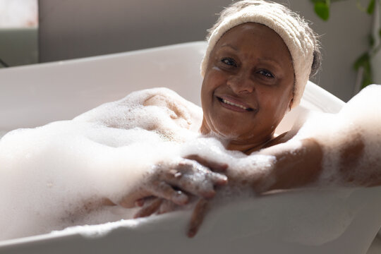 Senior African American woman reclining in bathroom tub with foam, wearing terry headband, smiling
