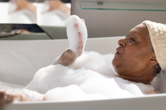 Senior African American woman reclining in tub soaking in foam towel turban mirror shelf chrome tap