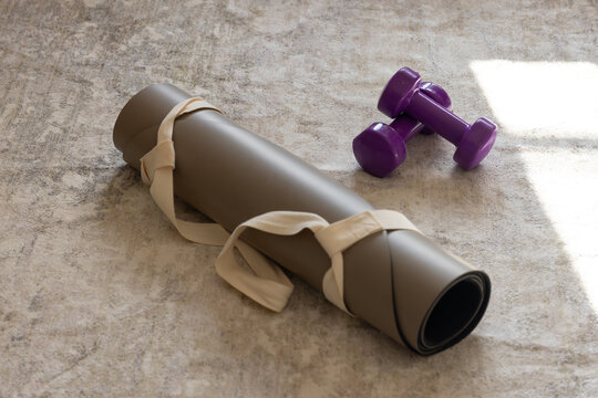 Rolled gray yoga mat is lying on beige carpet, strap looping, purple dumbbells resting in sunlight