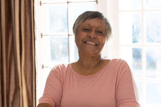 Senior woman smiling sitting at home by window wearing light pink longsleeve top with curtains