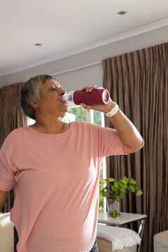 Senior woman standing in living room drinking from bottle with red sleeve wearing white tracker