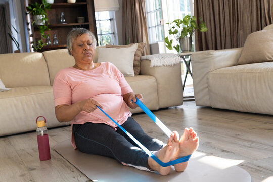 Senior African woman in pink tee sitting on mat pulling blue strap near bottle at home