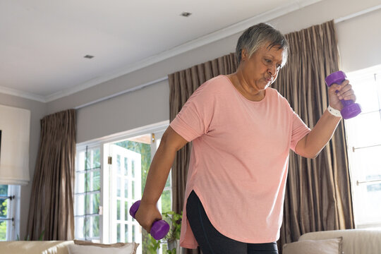 Senior African American woman in pink top exercising with purple weights in living room, copy space