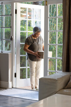 Mature woman standing at home in white French doors holding cardboard package, wearing olive top