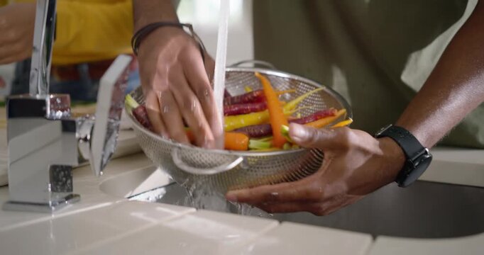 Couple, man holding colander rinsing dirty carrots under running faucet, woman in yellow watching