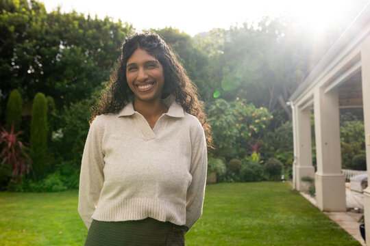 Adult Indian woman standing and smiling on well-kept lawn near patio, wearing cream sweater