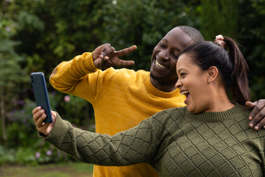 African American couple posing in garden wearing knits woman holding blue smartphone laughing