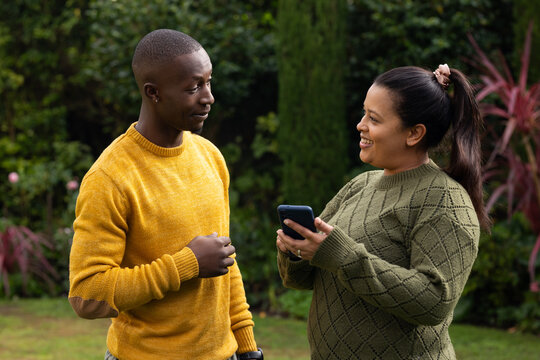 Couple standing on lawn, man in mustard sweater with watch, woman holding smartphone in olive knit