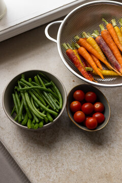 Colorful carrots resting in colander, green beans, cherry tomatoes on speckled beige top by window