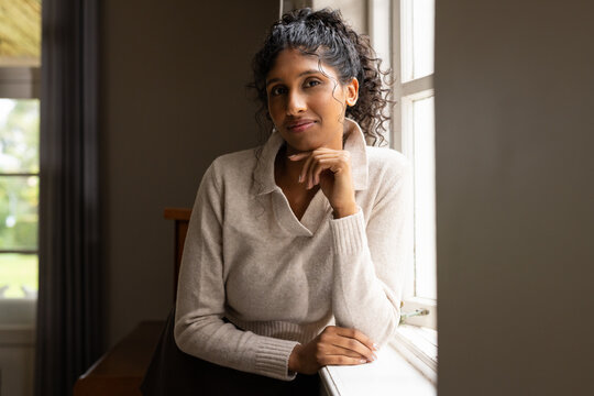 African American woman in beige sweater sitting at home by window leaning on sill touching chin