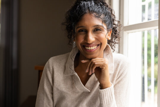 African American woman smiling, resting chin on right-hand at window at home in beige sweater