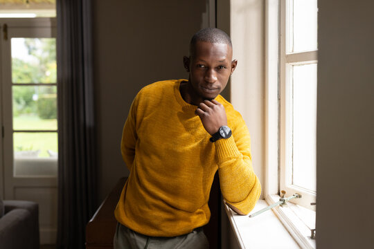 African American man leaning on white window, resting chin, wearing mustard sweater and wristwatch