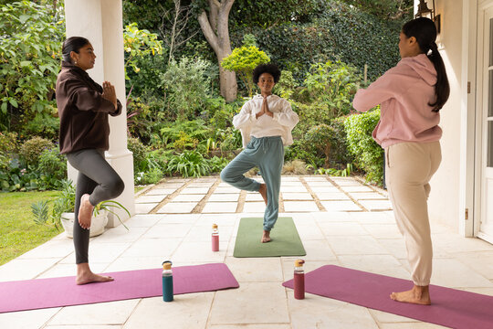 Diverse female friends practicing tree pose on covered home patio with mats and water bottles