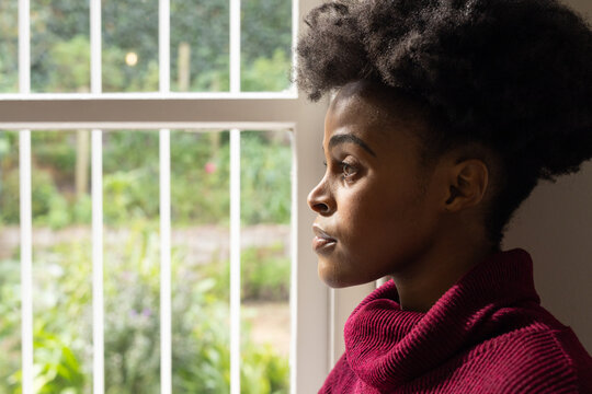 African adult woman gazing out white window with vertical bars in maroon ribbed sweater, copy space