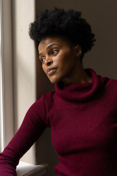Adult African woman leaning on sill at corner in ribbed burgundy turtleneck bathing in sidelight