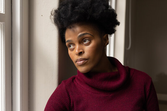 African-American woman leaning on wall by window with blind cord, in maroon sweater gazing upward