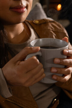 African American woman holding ridged gray mug near mouth on couch, wearing tan jacket
