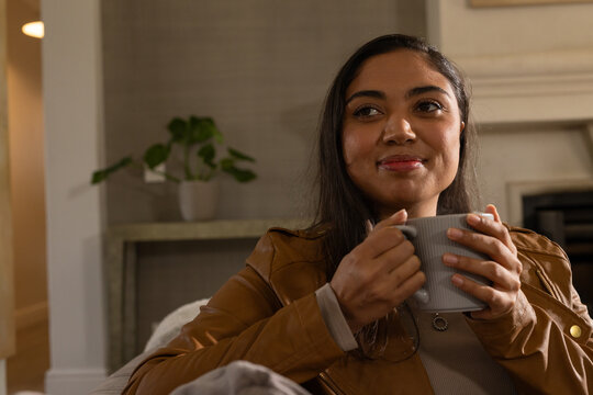 Adult African American woman wearing brown leather jacket holding ribbed mug near chest on sofa