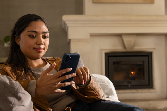 African American woman wearing leather jacket sitting on sofa holding smartphone by fireplace