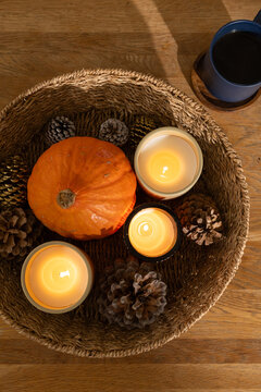 Round wicker basket holding small orange pumpkin, lit glass candles, pine cones on wooden table