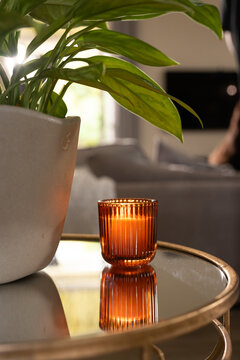 Amber ribbed glass candle is glowing on round glass-top table with brass rim, plant backlit