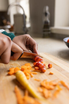 Adult female green-knit sleeve slicing tricolor carrots using chef-knife on wood-board at sink