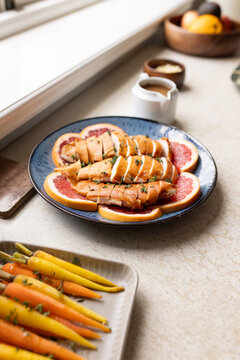 Blue ceramic plate is holding glazed protein slices over grapefruit on countertop with carrots