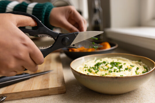 Woman wearing green striped sweater snipping chives with black shears over mashed potatoes