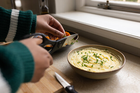 Indian woman in green sweater trimming herbs with shears beside mashed potato by window, copy space