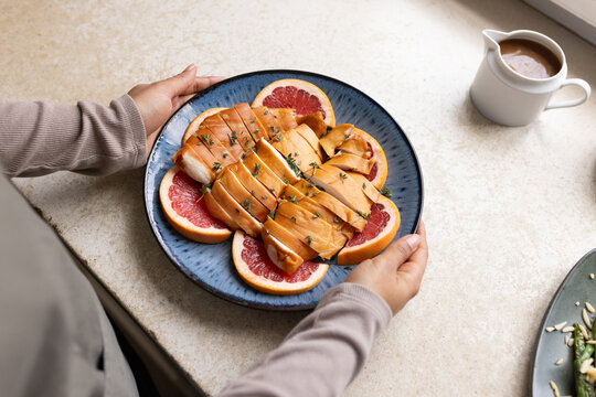 Adult woman in ribbed-top steadying blue plate with glazed fish on citrus halves at kitchen counter