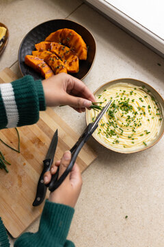 Adult female in green sweater cutting chives with shears over mashed potatoes on kitchen counter