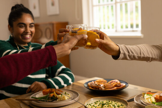 Diverse three adult women sitting at wooden dining table, raising glasses with orange slices