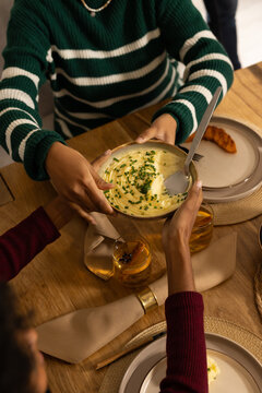 African American and Indian women passing ceramic mashed-potato bowl with chives at dining table