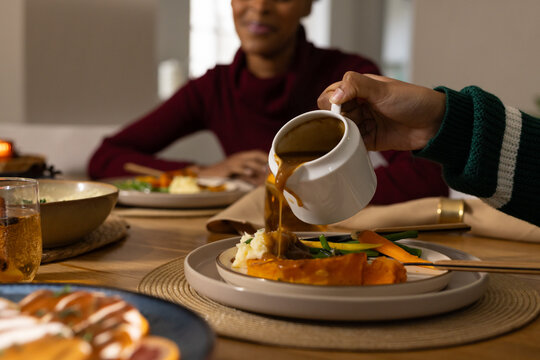 Diverse female companions dining at home, striped sweater pouring gravy from white jug onto plate