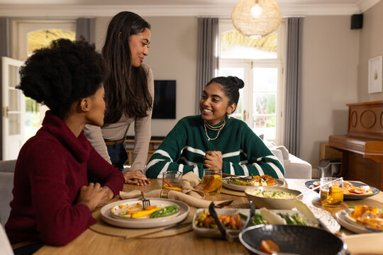 Diverse female friends sitting and chatting around wooden dining table at home with plated meals