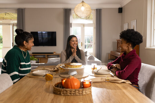 Diverse female friends sitting around wooden dining table at home, sharing cake and pumpkin tray
