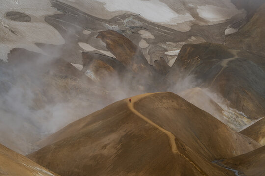 View of a person in a red jacket walking along a narrow mountain ridge surrounded by geothermal steam and colorful rhyolite peaks in Iceland.