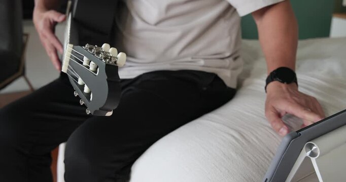 Senior Chinese man tuning black nylon guitar using tablet tuner cues in bedroom, preparing to play