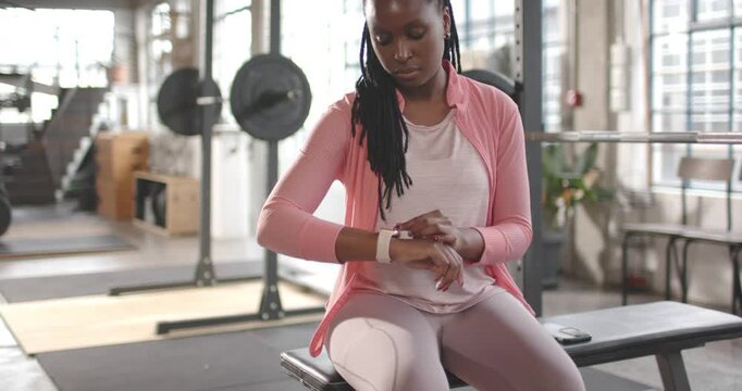 African American woman in pink jacket on bench tapping smartwatch, prepping for workout in loft gym
