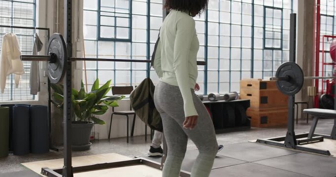 Diverse training pair entering gym carrying duffel, walking toward squat rack preparing for workout