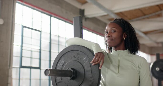 Mid adult female noticing camera, leaning on loaded barbell at gym, smiling in green zipup top
