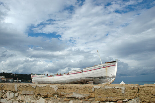 An old white wooden fishing boat pulled ashore on a coastal stone wall under a dramatic blue sky filled with white and grey clouds.