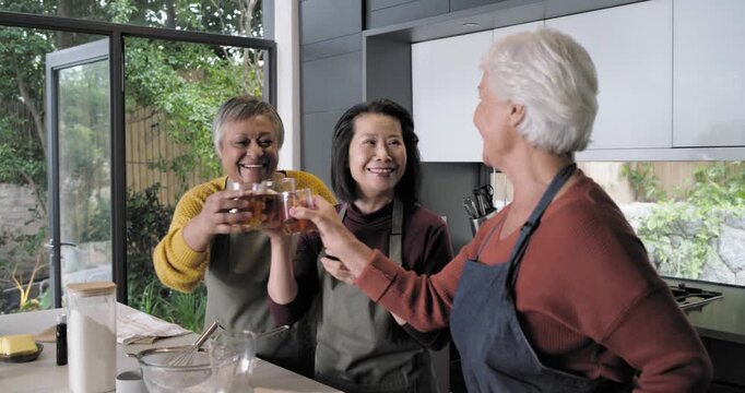 Diverse women, senior woman raising glass, clinking, celebrating, mixing bowl at kitchen island
