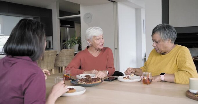Diverse women sharing pie at home table, center senior starting story, catching up, phone on table