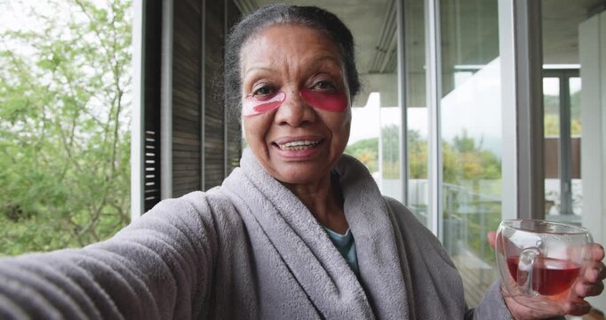 Senior African-American woman holding phone showing pink eye patches while sipping tea on balcony