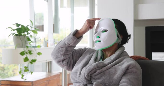 Senior African American at home wearing robe raising hand fitting LED mask activating green glow