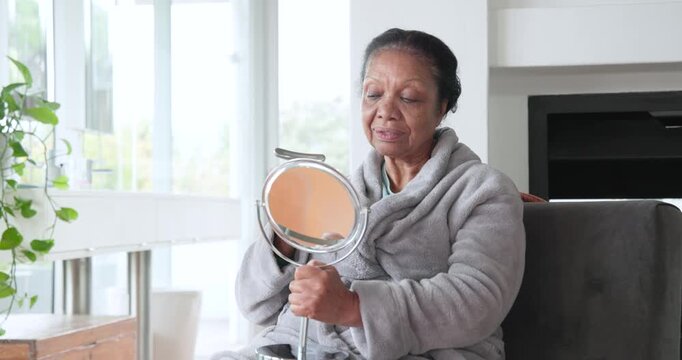 Senior African woman in robe with mirror and eye tool grooming eyes, checking alignment at home