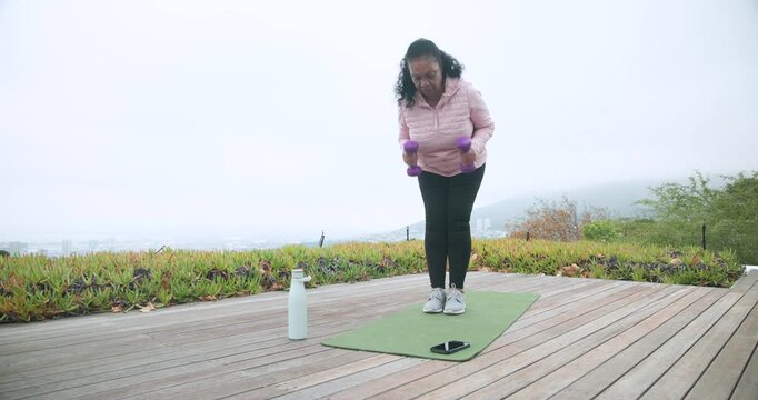 Senior African woman starting workout on green mat on deck, lifting purple dumbbells for fitness